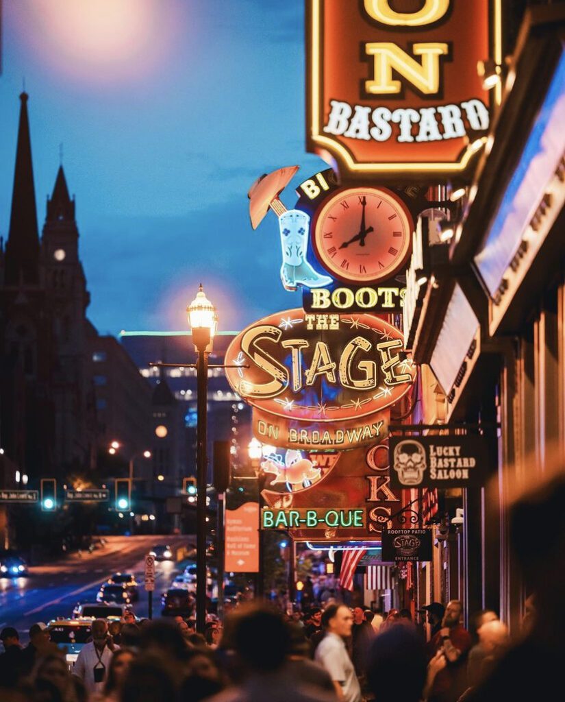 Exterior of The Stage on Broadway in Nashville, with neon guitar signs and crowds gathering outside the honky-tonk.