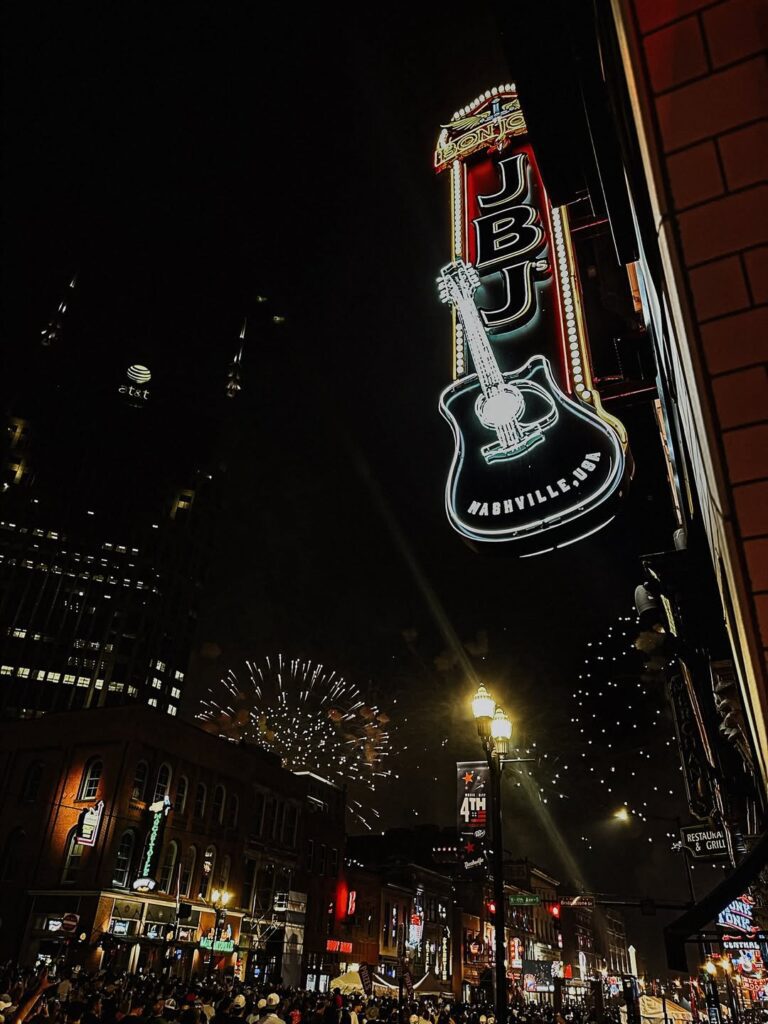 JBJ's neon guitar sign on Nashville's Broadway at night with fireworks and crowds below.